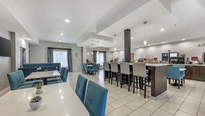 Un espace de restauration moderne dans un hôtel, avec des sièges bleus rembourrés, un îlot central doté d’une machine à café et une cuisine en arrière-plan.