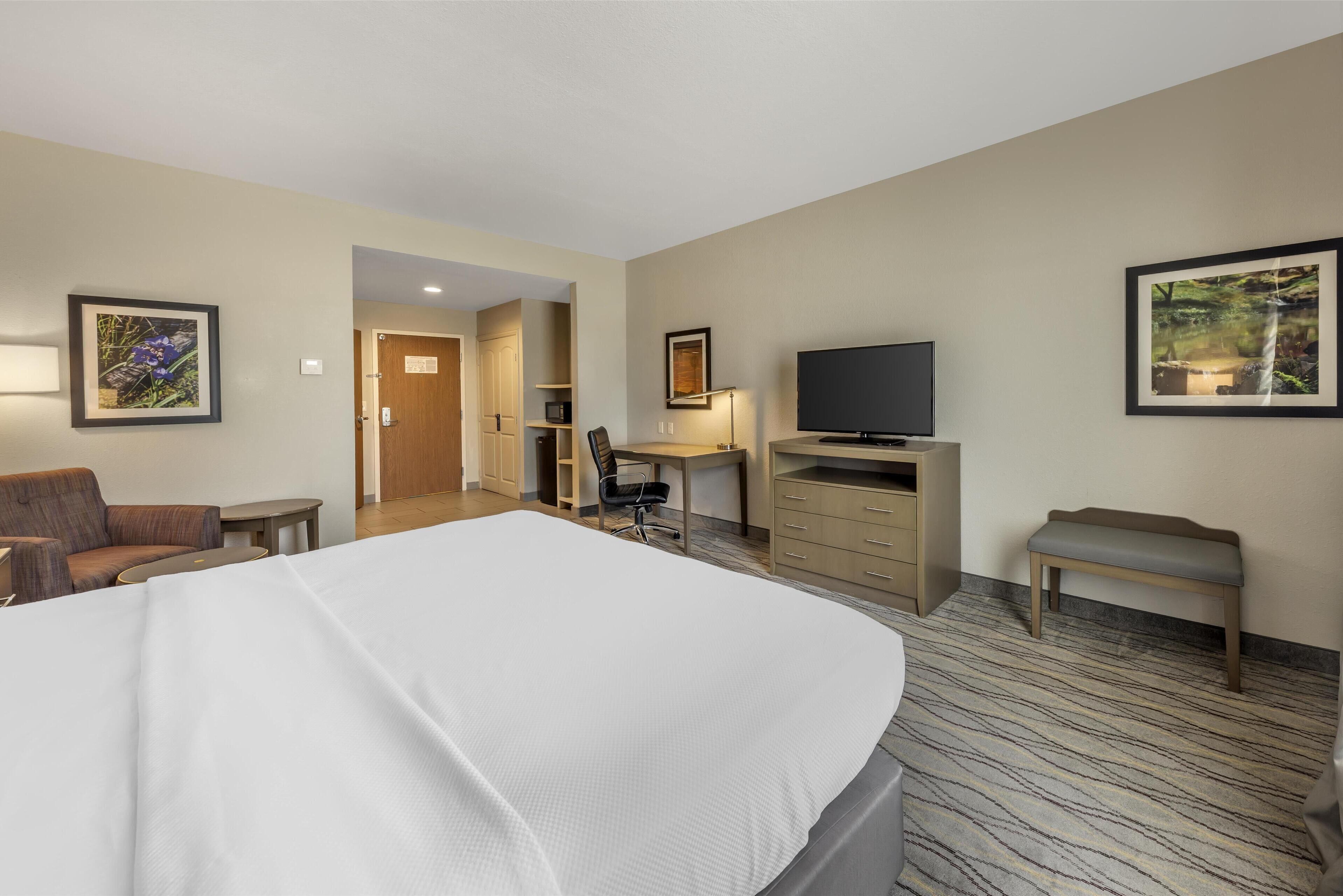 A hotel room with a large bed, a desk, a television, a chair, and a bench.