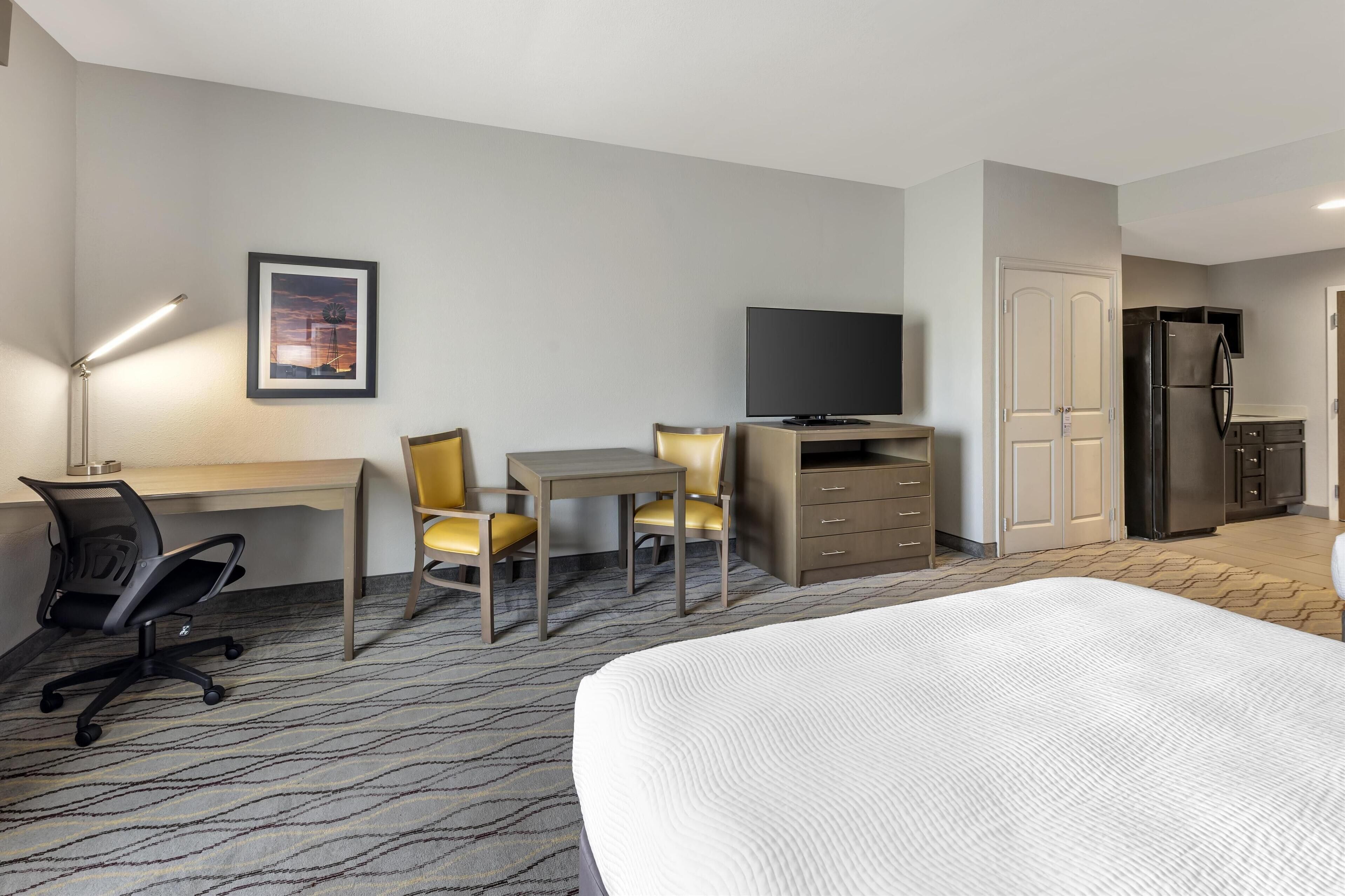 A hotel room with a bed, desk, chairs, a television, a refrigerator, and a wardrobe.