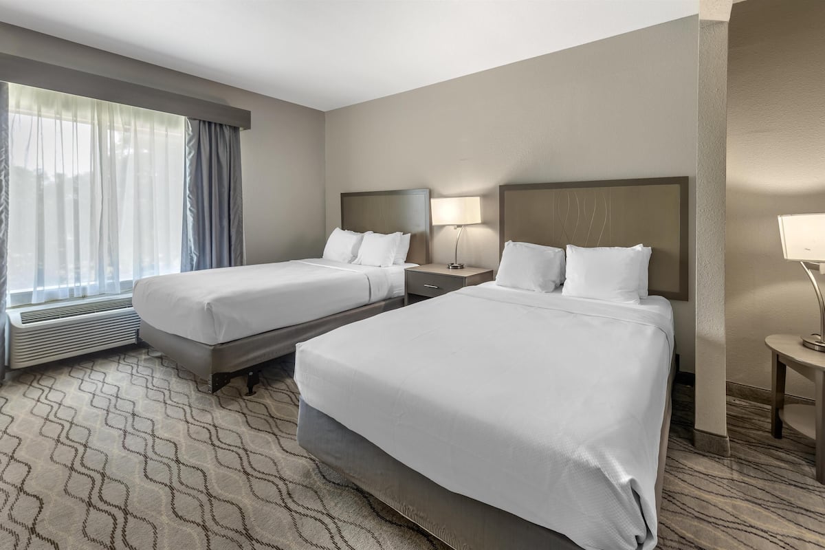 A hotel room with two beds, a large window with curtains, a flat-screen TV, and a small table with a lamp.