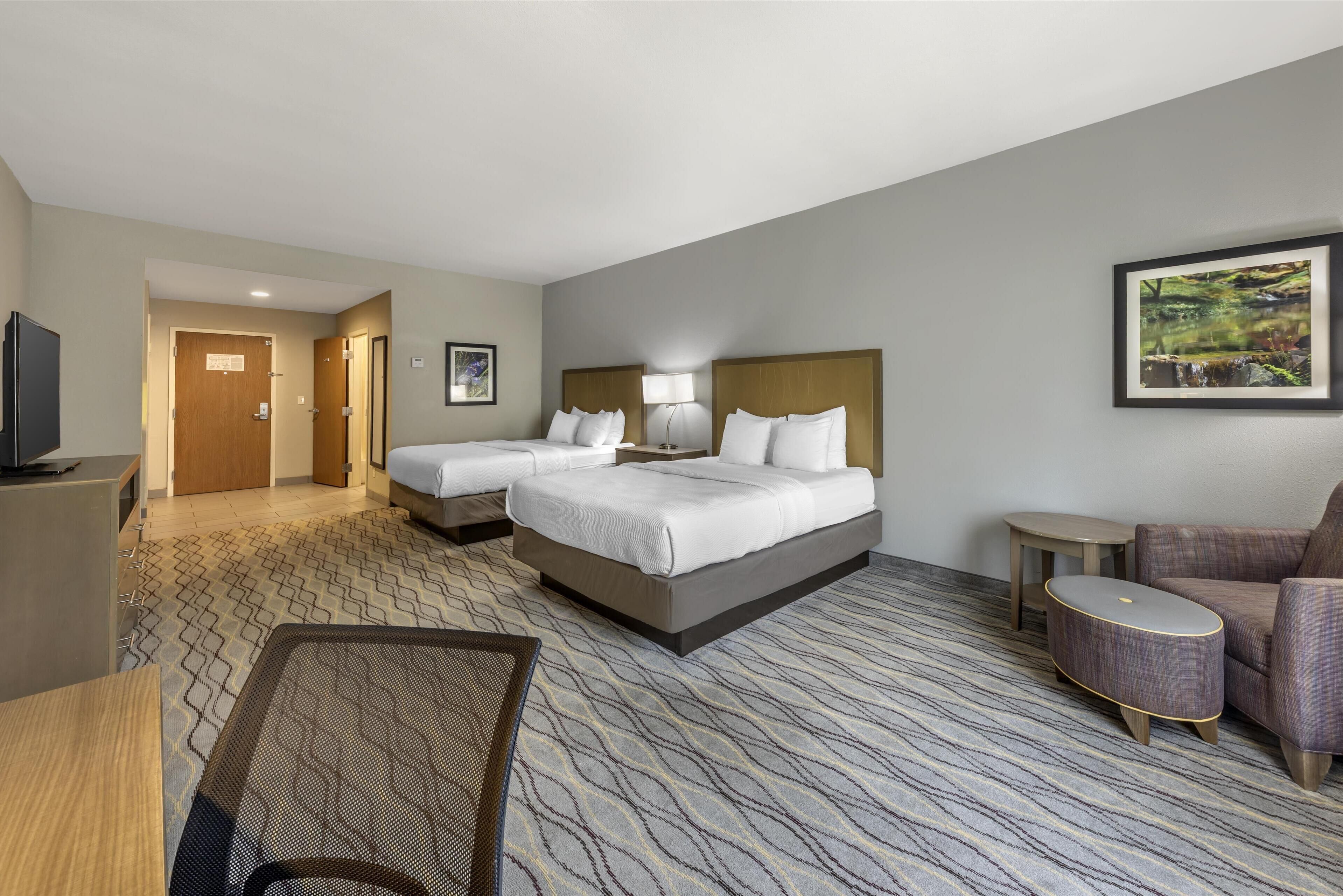 A hotel room with two beds, a desk, a chair, a small table, and a painting on the wall.