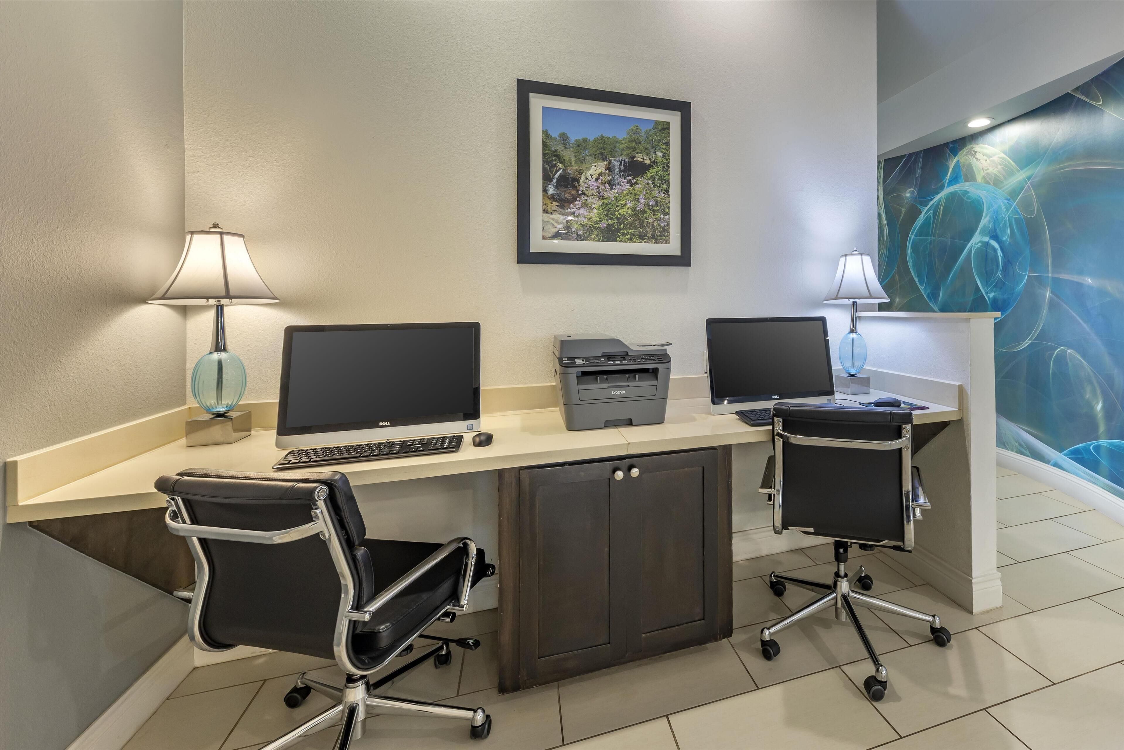 Un espacio de oficina moderno con dos escritorios, cada uno con un monitor de computadora, teclado e impresora. Hay dos sillas de oficina, un cuadro en la pared y un elemento decorativo en la pared.