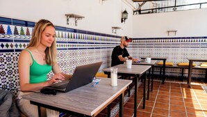 A person using a laptop at a table in a room with patterned tile walls and a tiled floor. - Triana Backpackers (Seville)