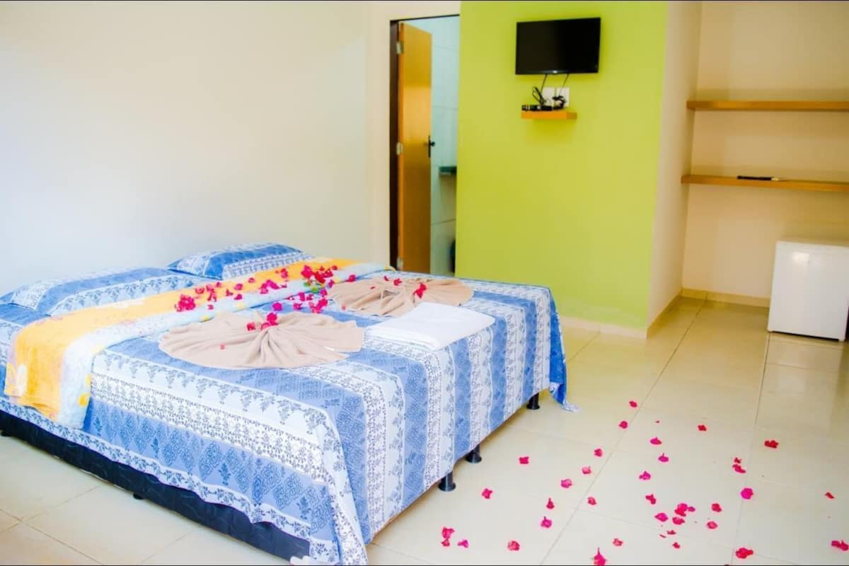 A hotel room with a bed covered in blue and white patterned bedding, a yellow throw blanket, and pink flower petals scattered on the floor.