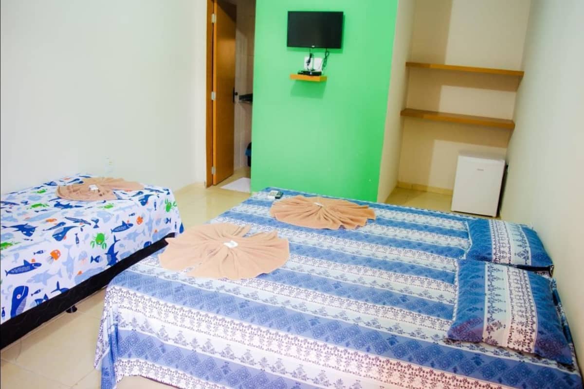 A room with two beds, one with a blue and white patterned bedspread and the other with a blue and white striped bedspread. There is a green wall with a mounted television and a small shelf.