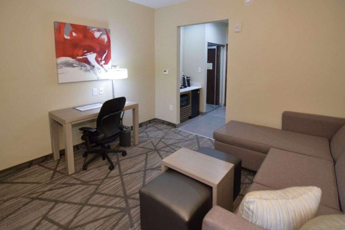 Habitación de hotel con escritorio, silla de oficina, sofá y reposapiés. Hay un cuadro en la pared y una puerta que da a una kitchenette.