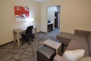 A hotel room with a desk, office chair, sofa, and ottoman. There is a painting on the wall and a door leading to a kitchenette. - Comfort Inn & Suites Houston I-45 North - IAH (Houston)