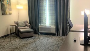 A hotel room with a sofa, a standing lamp, a large window with curtains, an air conditioning unit, and a desk with a computer. - Comfort Inn & Suites Houston I-45 North - IAH (Houston)