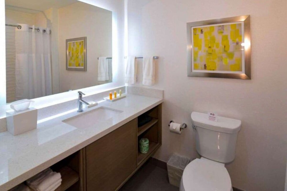 A bathroom with a white sink, mirror, and toilet. There are towels on the wall and a trash bin next to the toilet.