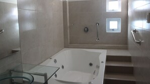 A modern bathroom with a glass-enclosed bathtub, a shower area with a handheld showerhead, and a set of stairs leading to a shower platform. - Hotel Emerawaa (Riohacha)