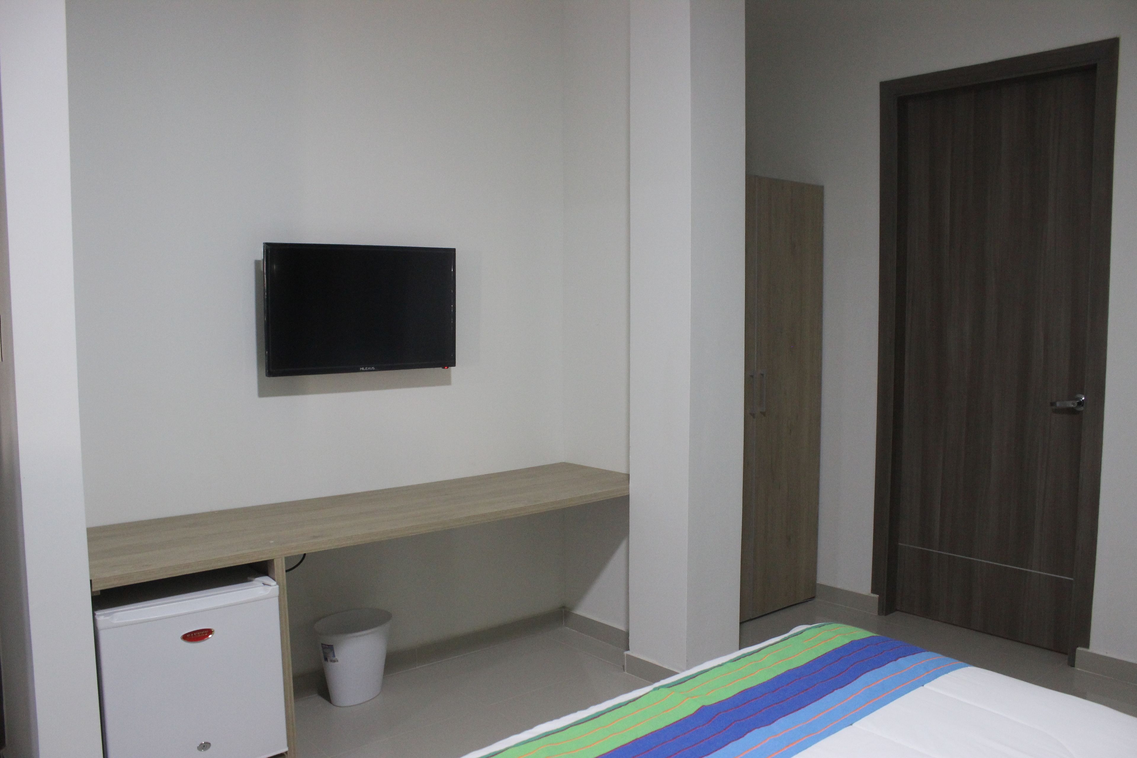 A bedroom with a bed, a television mounted on the wall, a small refrigerator, a wooden desk, and a door.