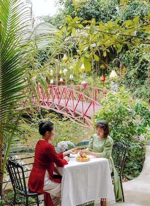 Two people dining at an outdoor table with a white tablecloth, surrounded by lush greenery and a pink bridge in the background. - Chez Loan Hotel (Nam Hoa Lu)