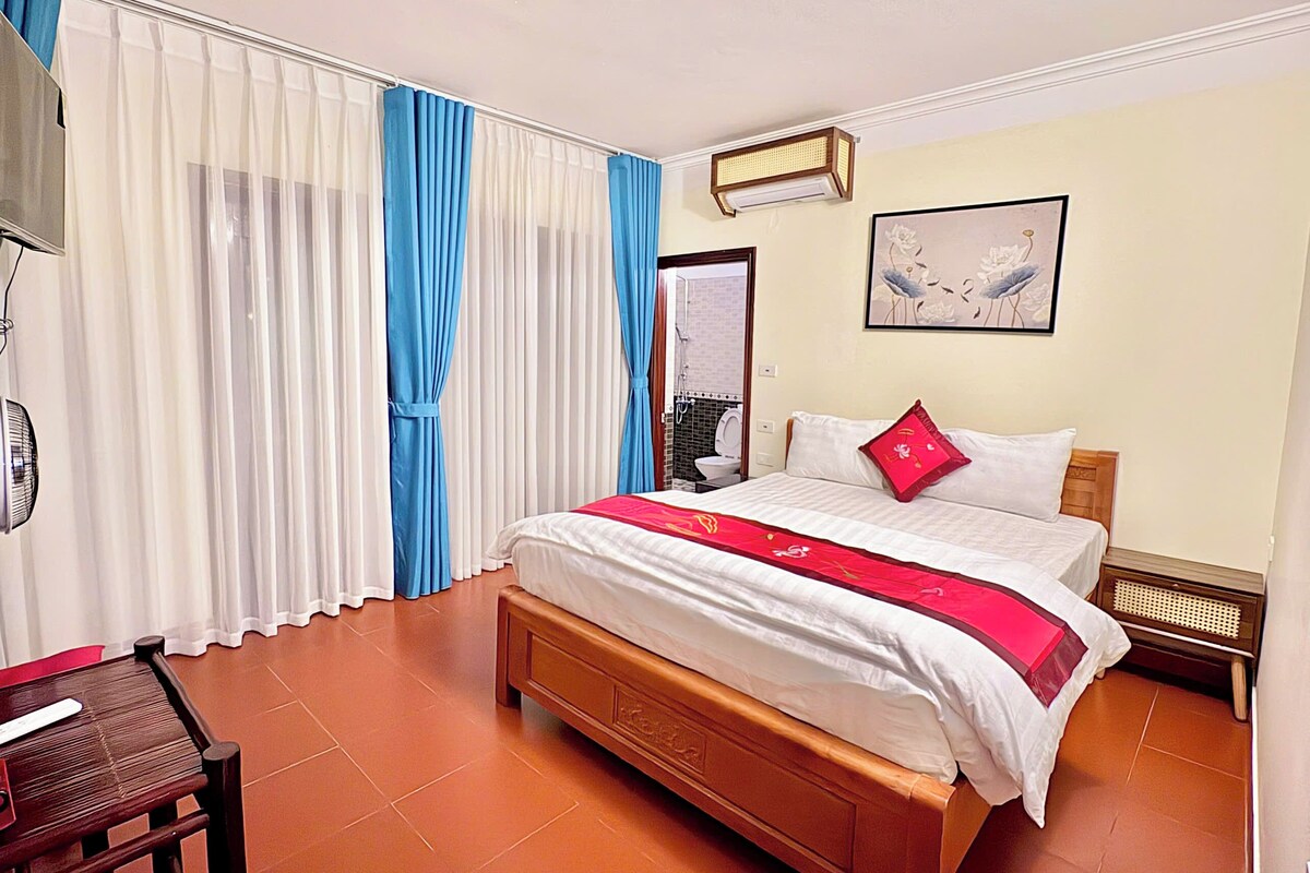 A hotel room with a large bed, a wooden nightstand, a desk with a chair, and a bathroom visible through a door.
