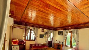 Una habitación con techo y paredes de madera, una cama con ropa blanca, una mesita, un banco y una ventana con cortinas.