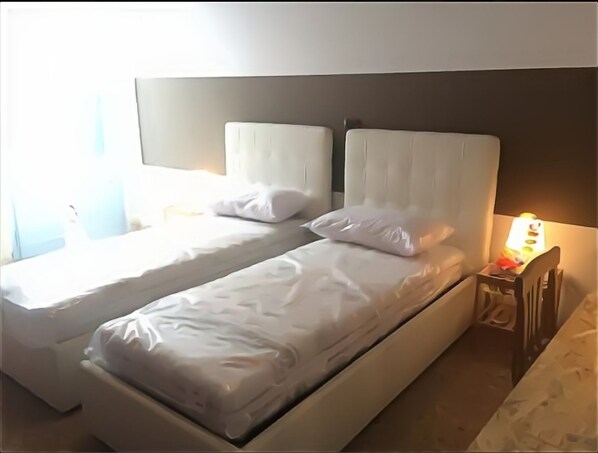 A twin bed with white bedding and headboards, a bedside table with a lamp, and a window with curtains.
