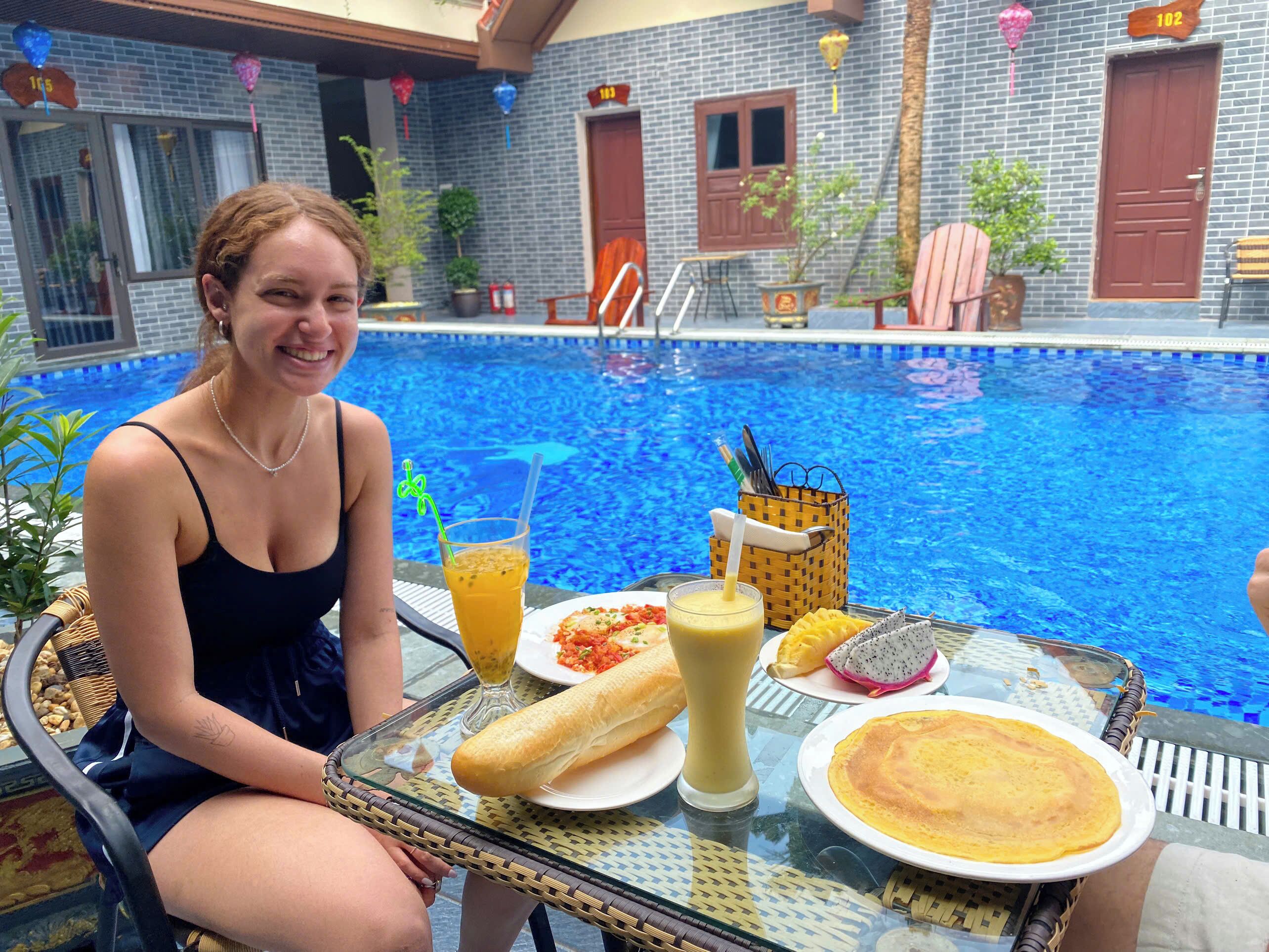 A person sitting at a table by a pool with a drink, a sandwich, and a plate of food.