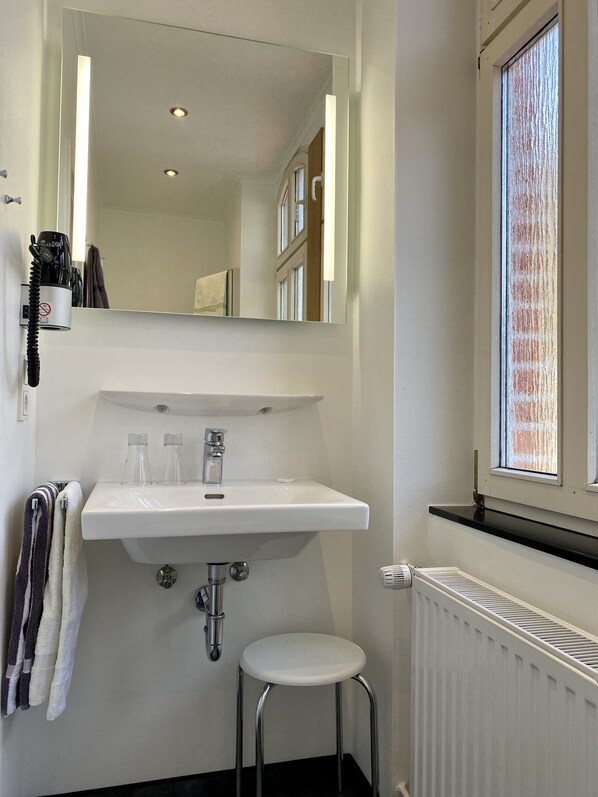A bathroom with a white sink, a mirror, a towel rack with towels, a radiator, and a window with blinds. - Haus Hohenzollern (Bad Bertrich)