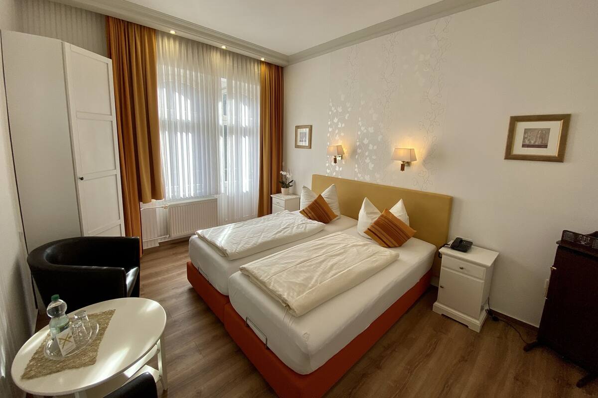 Quarto de hotel com duas camas, uma escrivaninha, uma cadeira e uma janela com cortinas.