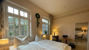 A hotel room with two beds, a desk, a chair, a lamp, and a view of the outdoors through large windows. - Haus Hohenzollern (Bad Bertrich)