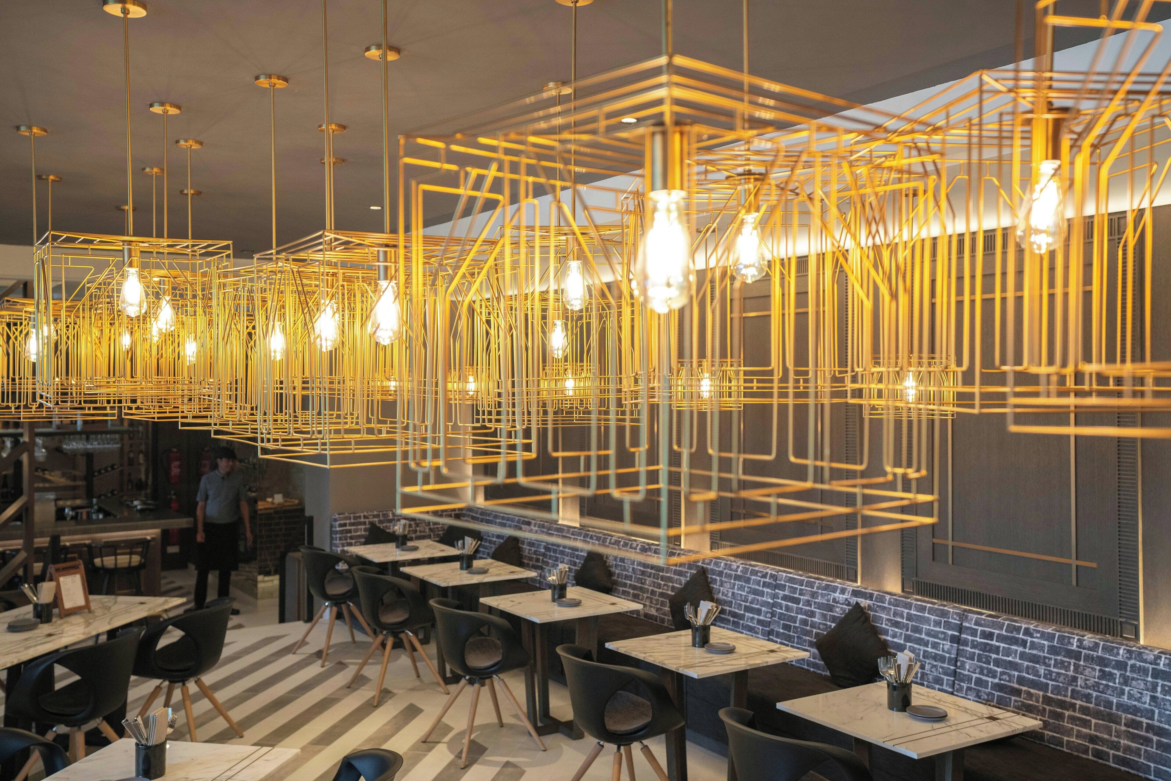 Interior de restaurante com mesas, cadeiras, luminárias pendentes e parede de tijolos.