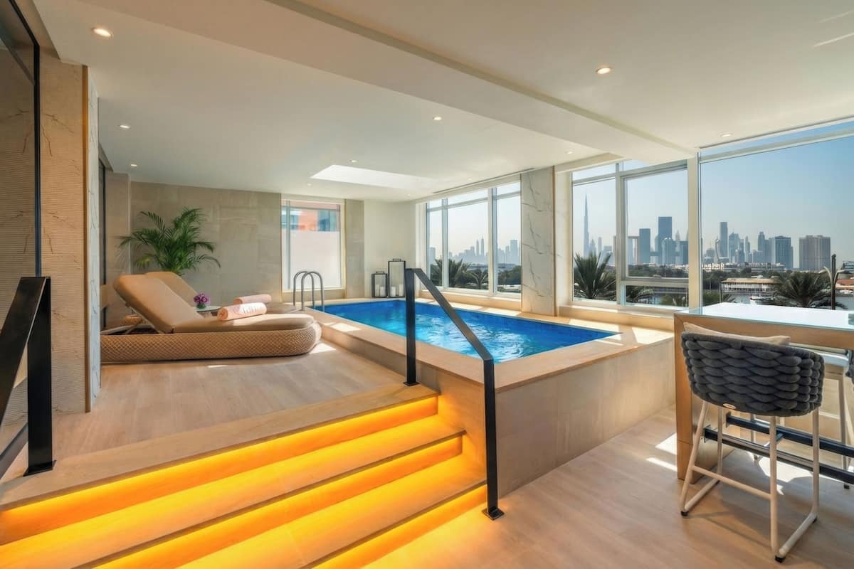Un espace piscine intérieur moderne offrant une vue sur les gratte-ciel de la ville, avec un fauteuil de relaxation et un escalier dont les marches sont illuminées.