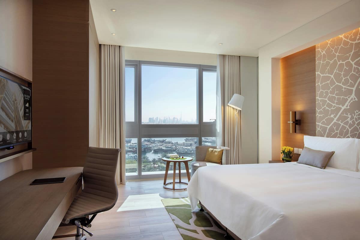 Une chambre d’hôtel avec un lit, un bureau, une chaise, une petite table et une vue sur le paysage urbain, visible à travers de grandes fenêtres.