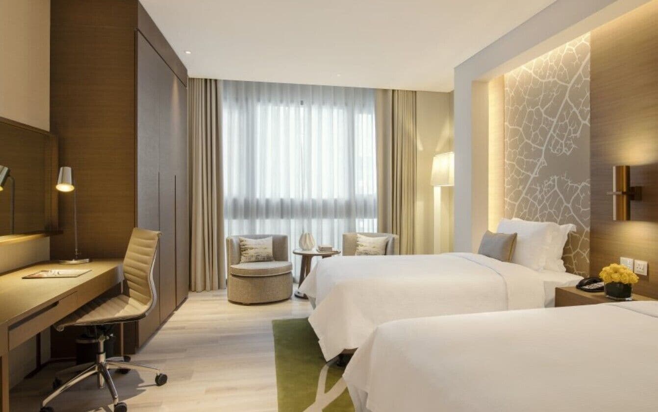 A hotel room with two beds, a desk, a chair, a small table, and a window with curtains.