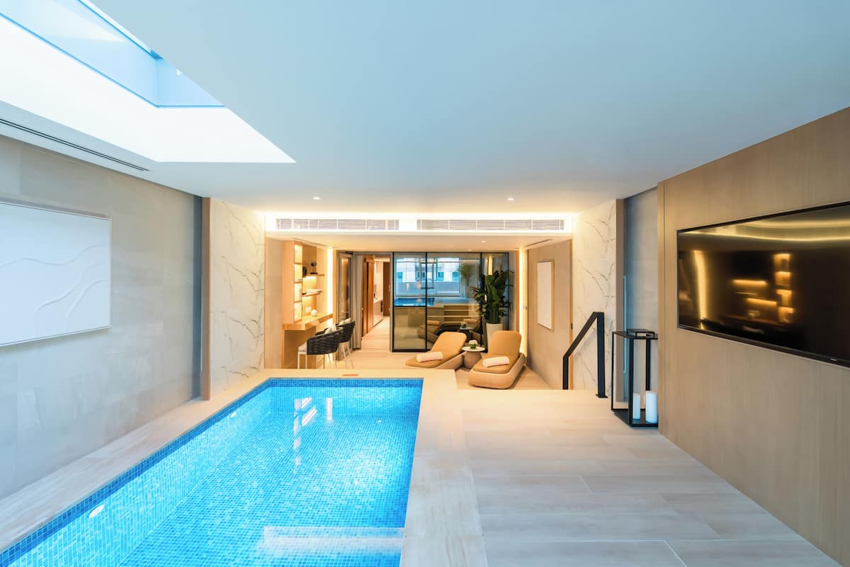 Une piscine intérieure moderne dotée d’un toit en verre, de chaises longues et offrant une vue sur l’extérieur.