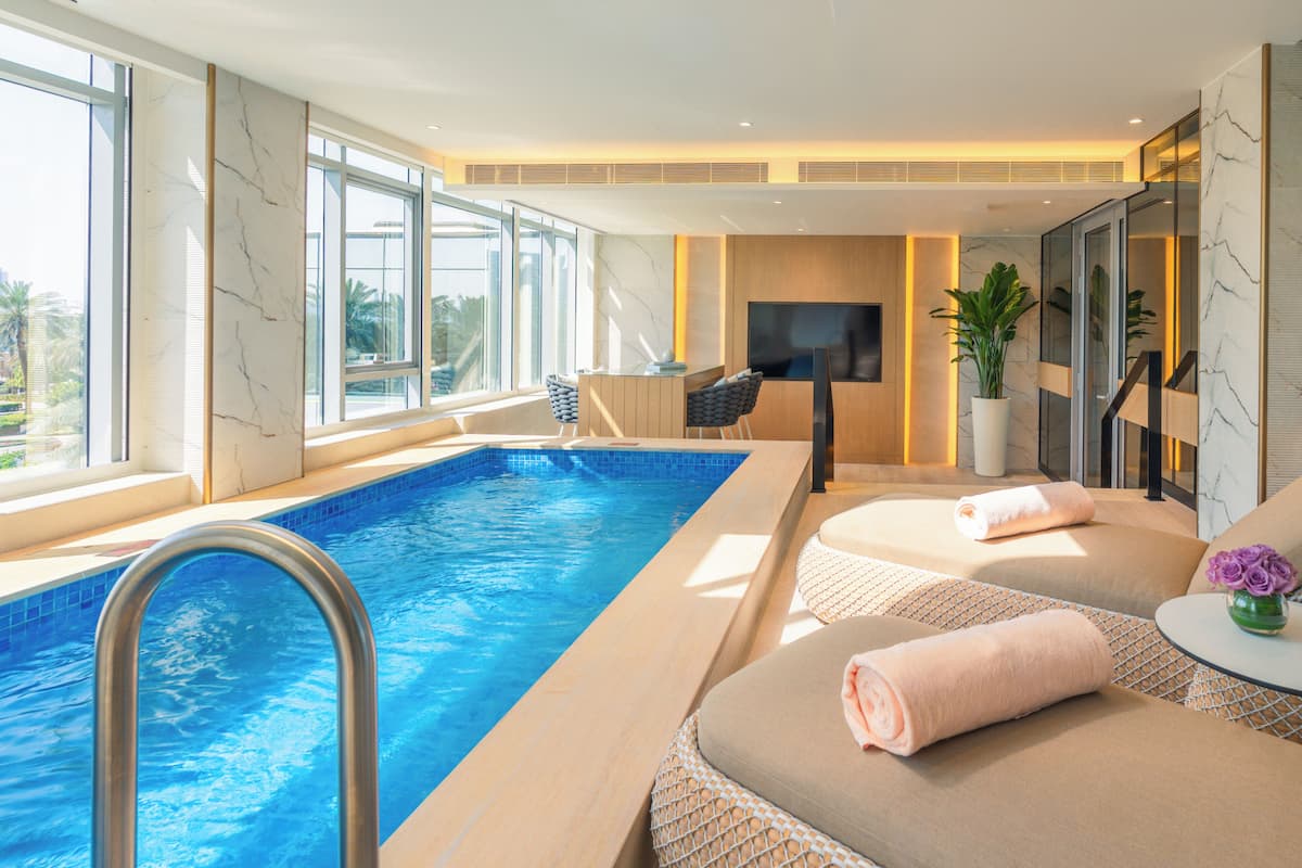 Une piscine donnant sur une pièce baignée de lumière, équipée d’un téléviseur, de chaises longues et d’une plante en pot.