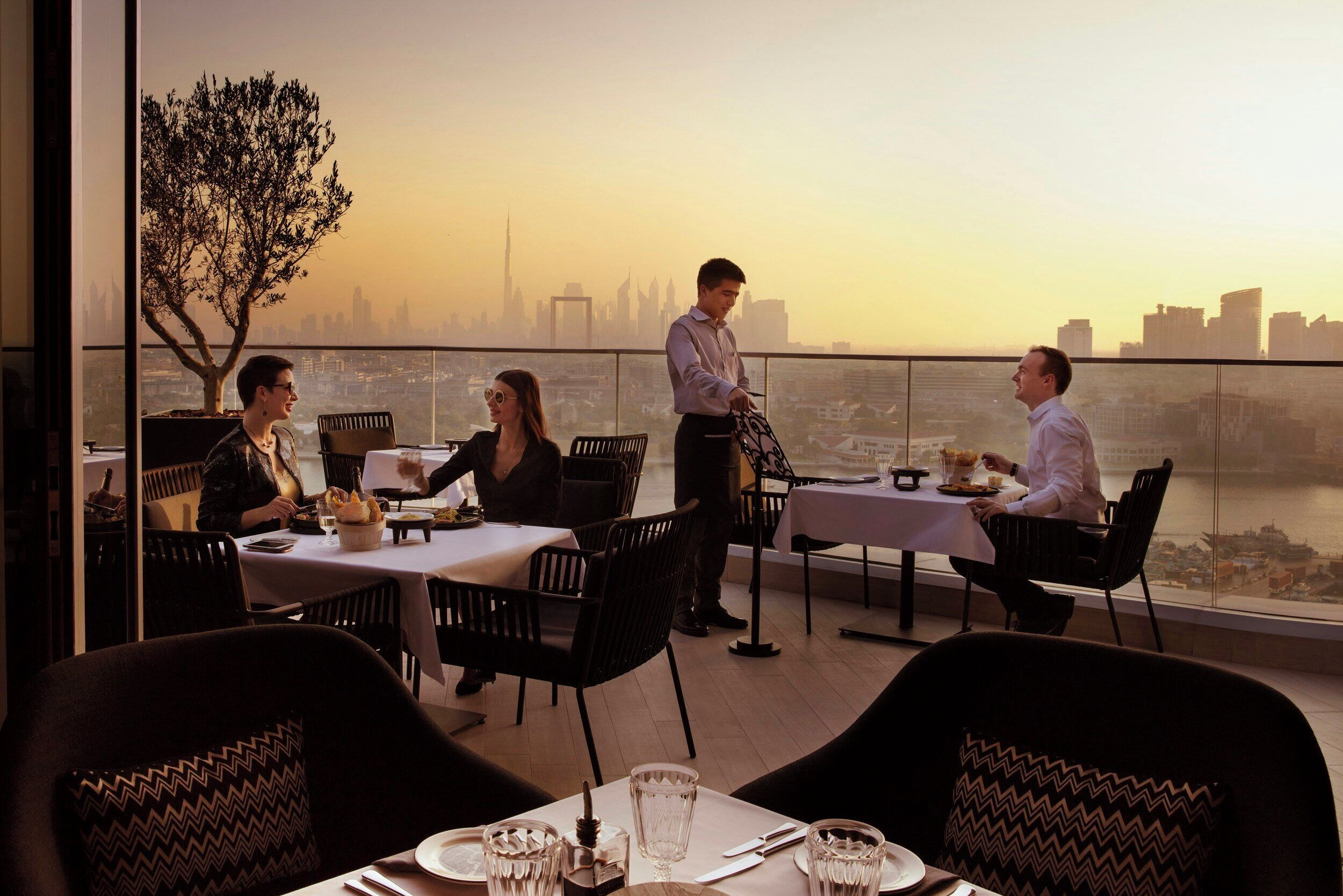 A restaurant with tables and chairs, a waiter, and a view of a city skyline.