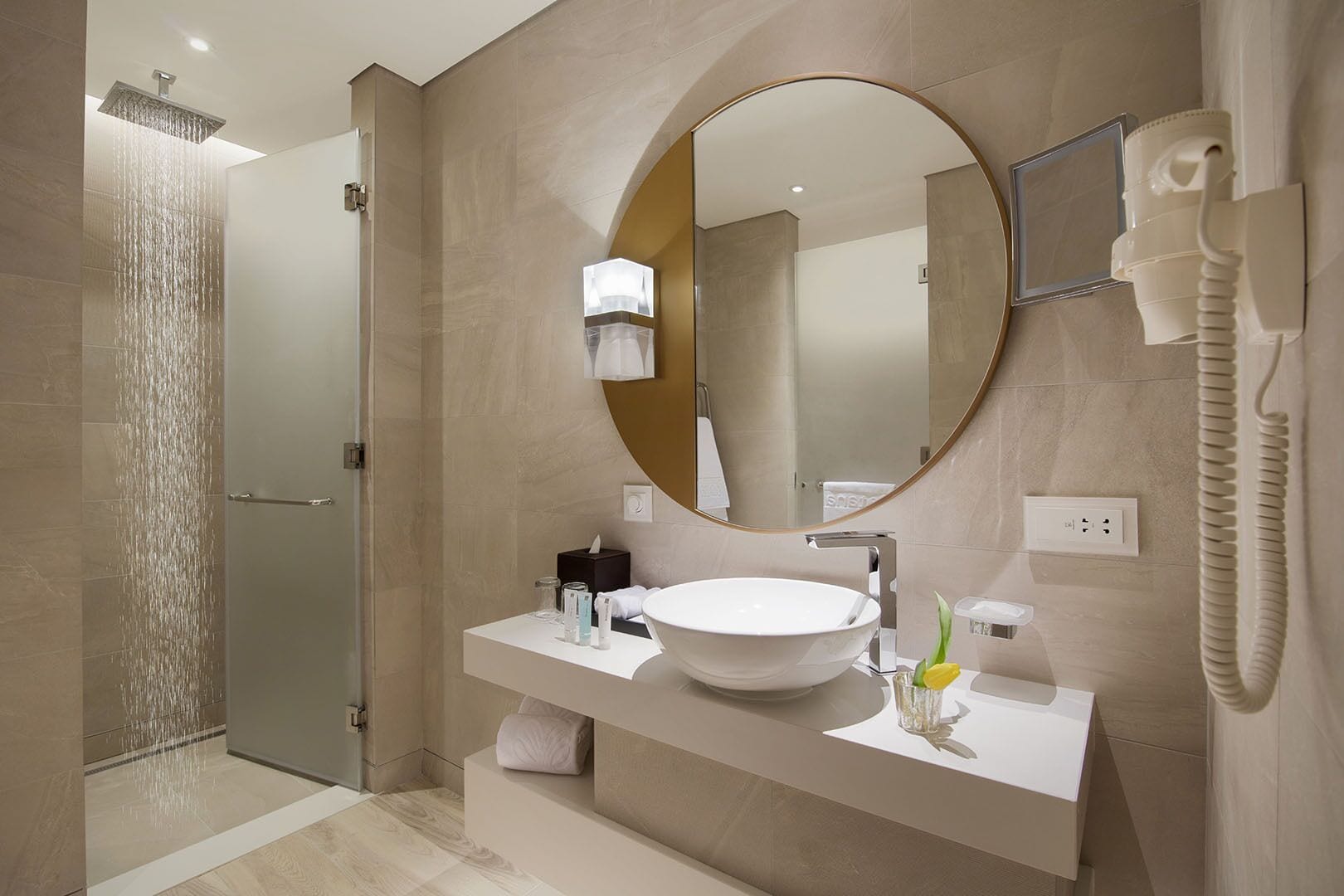 A modern bathroom with a glass-enclosed shower, a round mirror, a white sink, and a telephone.