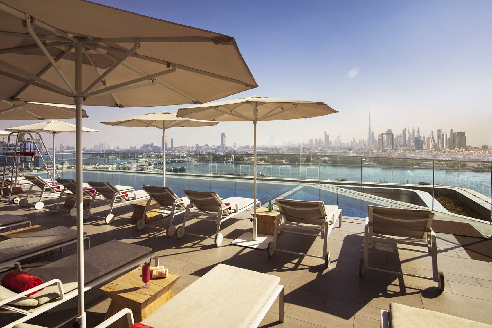 A rooftop terrace with lounge chairs, umbrellas, and a view of a city skyline and water body.