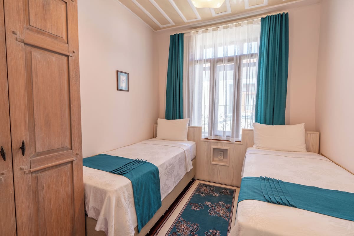 A room with two single beds, a wooden wardrobe, a window with curtains, and a small picture on the wall.