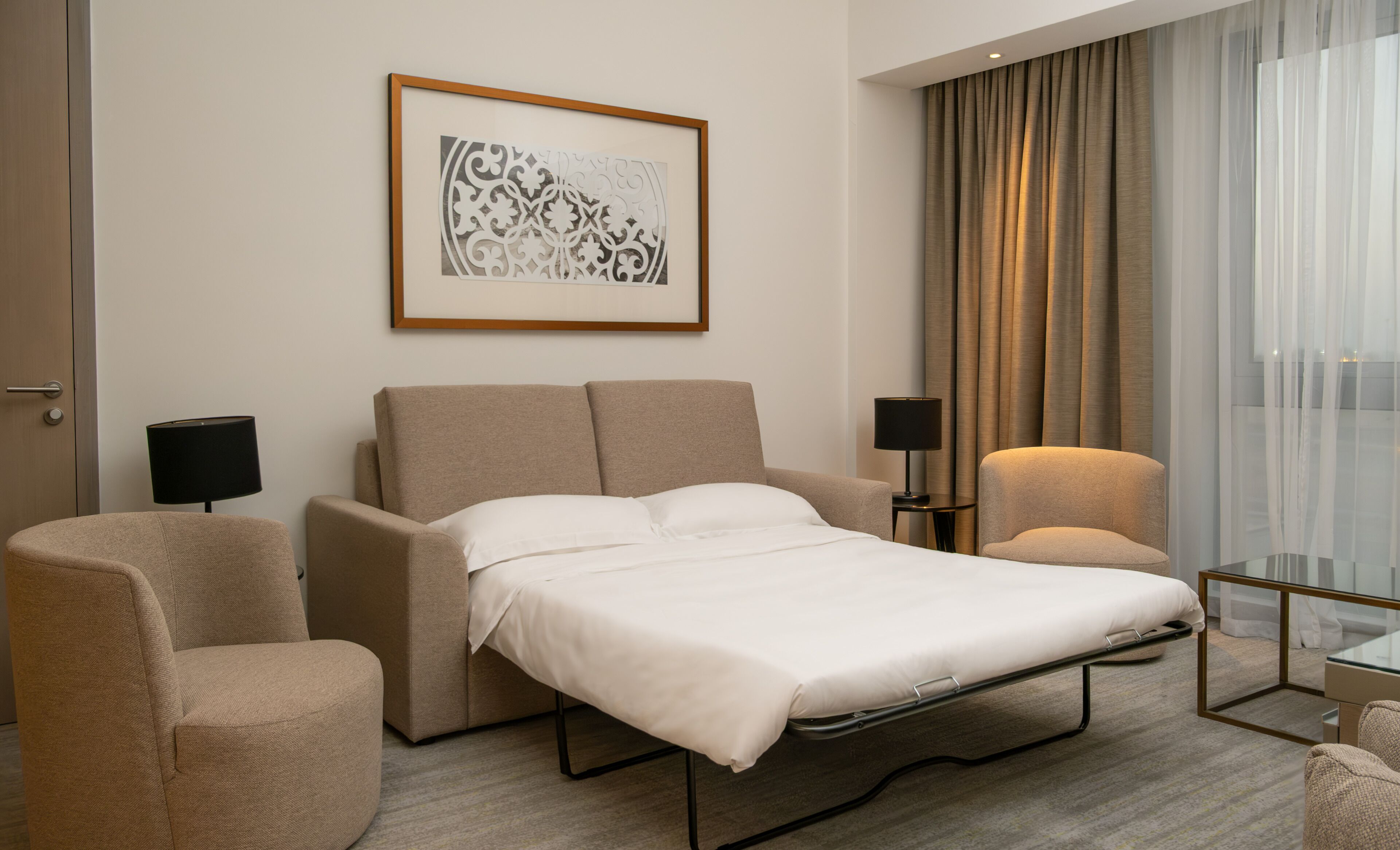 Una habitación de hotel moderna con una cama, dos sillones, una mesita y una obra de arte enmarcada en la pared.