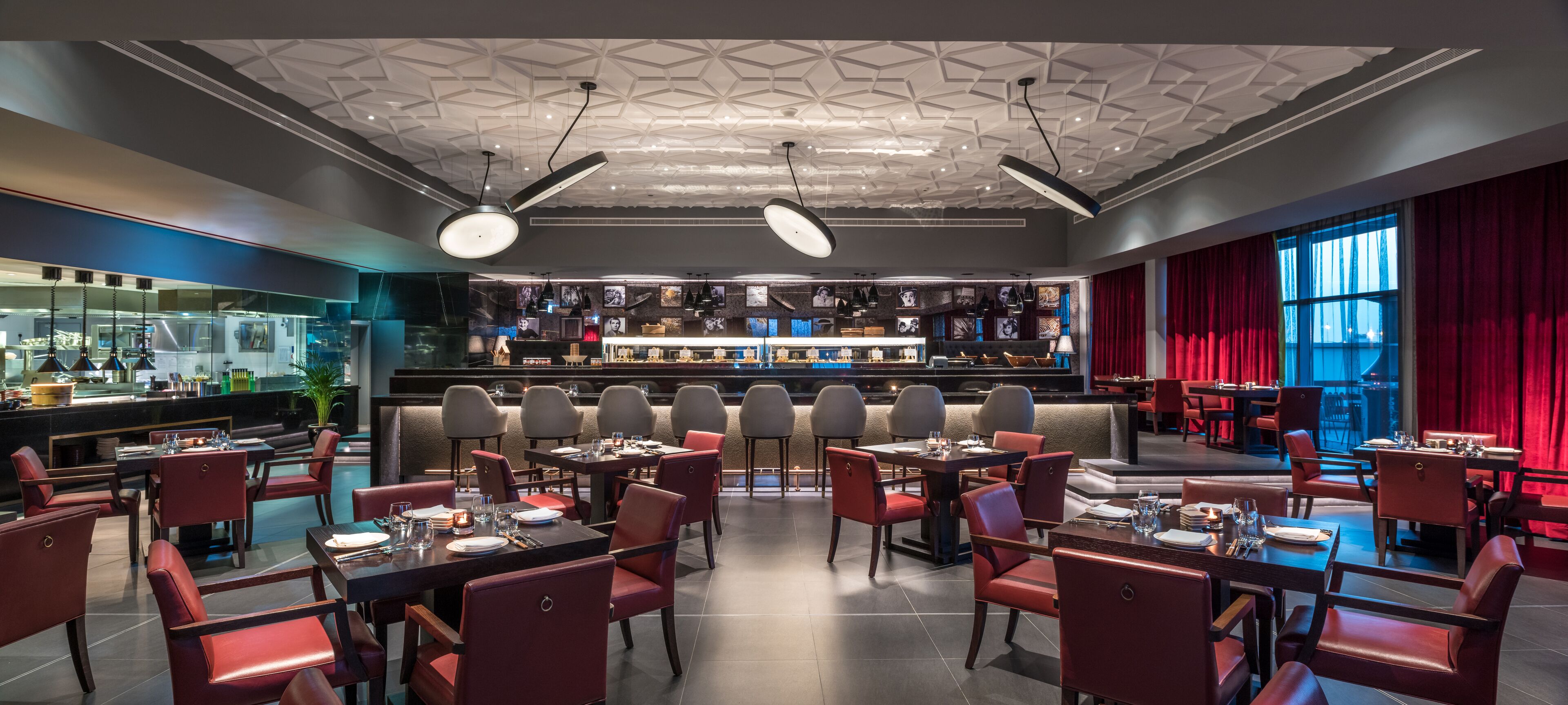 A modern restaurant with a bar area, red upholstered chairs, and a geometric ceiling design.