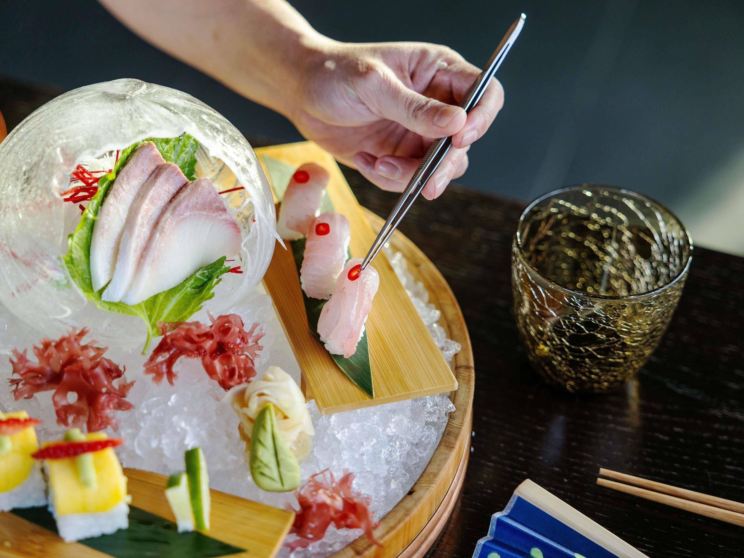Una persona usando palillos para tomar sushi de una bandeja de madera sobre una mesa.