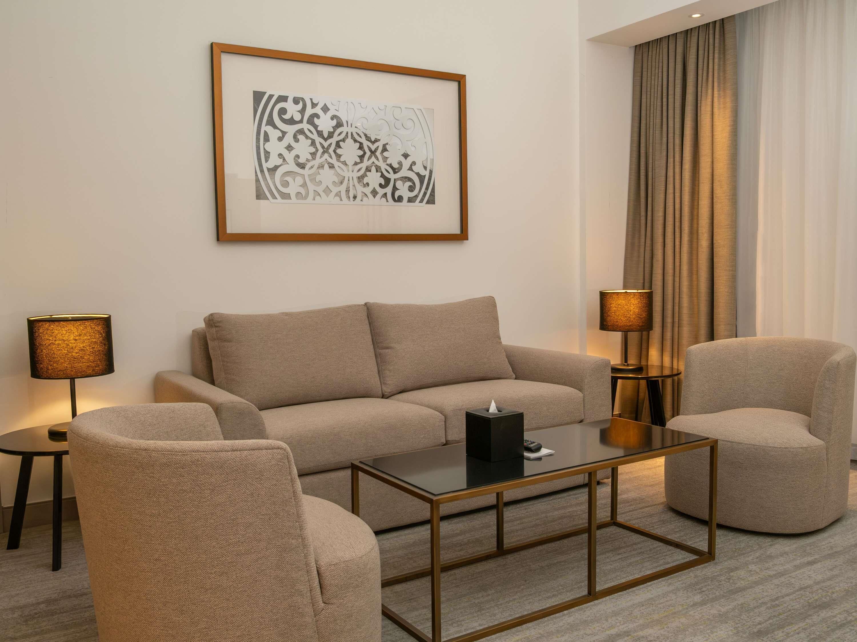 A modern living room with a sofa, two armchairs, a glass coffee table, and two lamps.