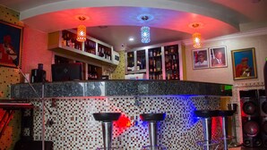 Un coin bar avec un mur recouvert de mosaïques, deux tabourets et une variété de bouteilles exposées sur des étagères.