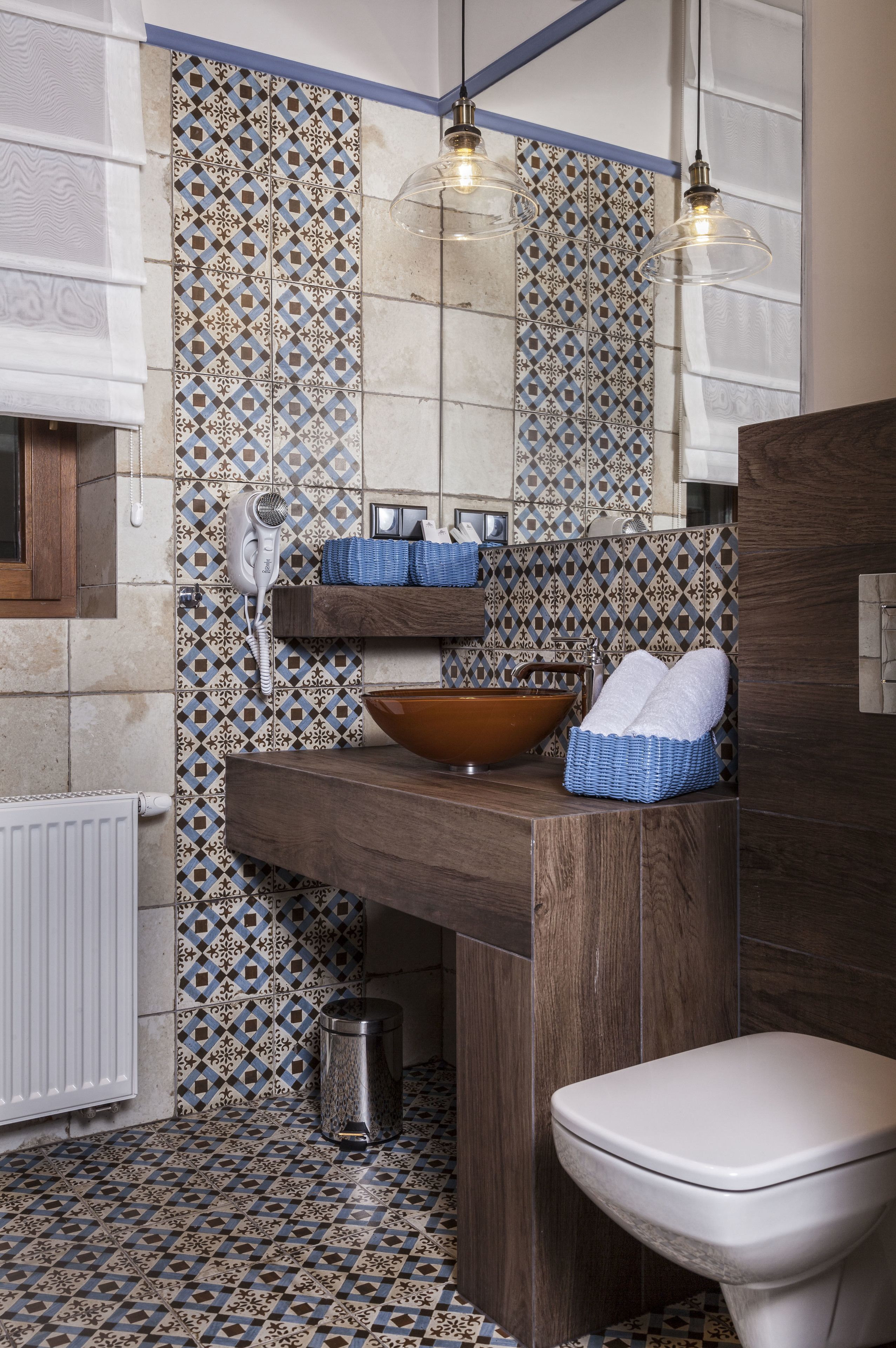 A bathroom with patterned tiles, a wooden vanity, a bowl sink, a toilet, and hanging pendant lights.