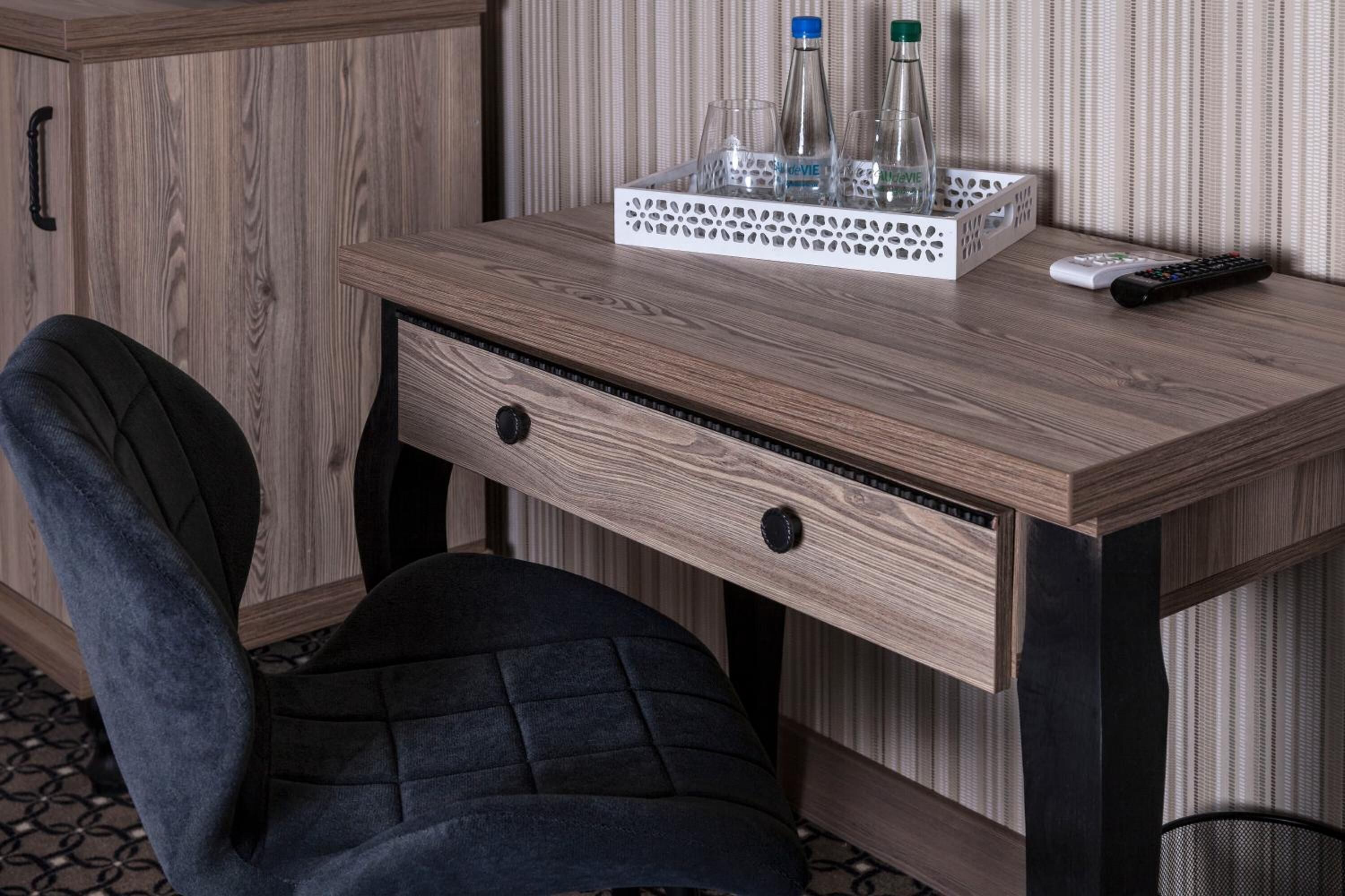 Un bureau en bois avec un plateau où sont posées des bouteilles et des verres, une télécommande et une chaise.