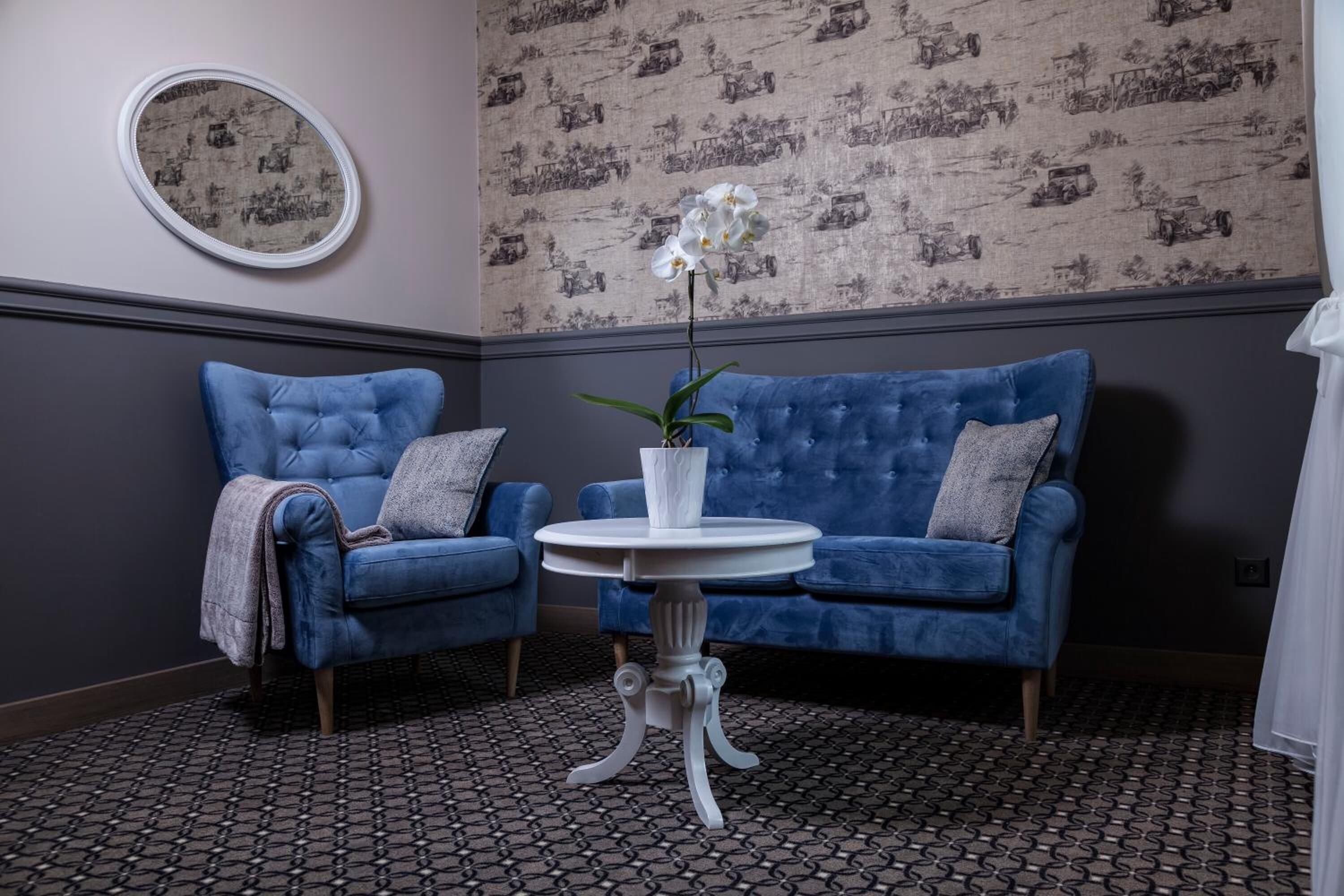 Una habitación con dos sillones azules, una pequeña mesa blanca con un jarrón de flores, papel tapiz estampado y un espejo redondo en la pared.