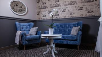 Una habitación con dos sillones azules, una pequeña mesa blanca con un jarrón de flores, papel tapiz estampado y un espejo redondo en la pared.
