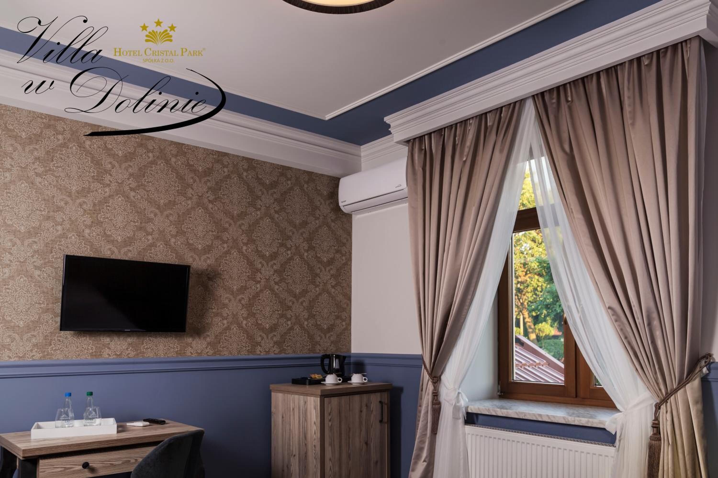 A hotel room with a patterned wallpaper, a flat-screen TV, a wooden cabinet, a window with curtains, and a radiator.