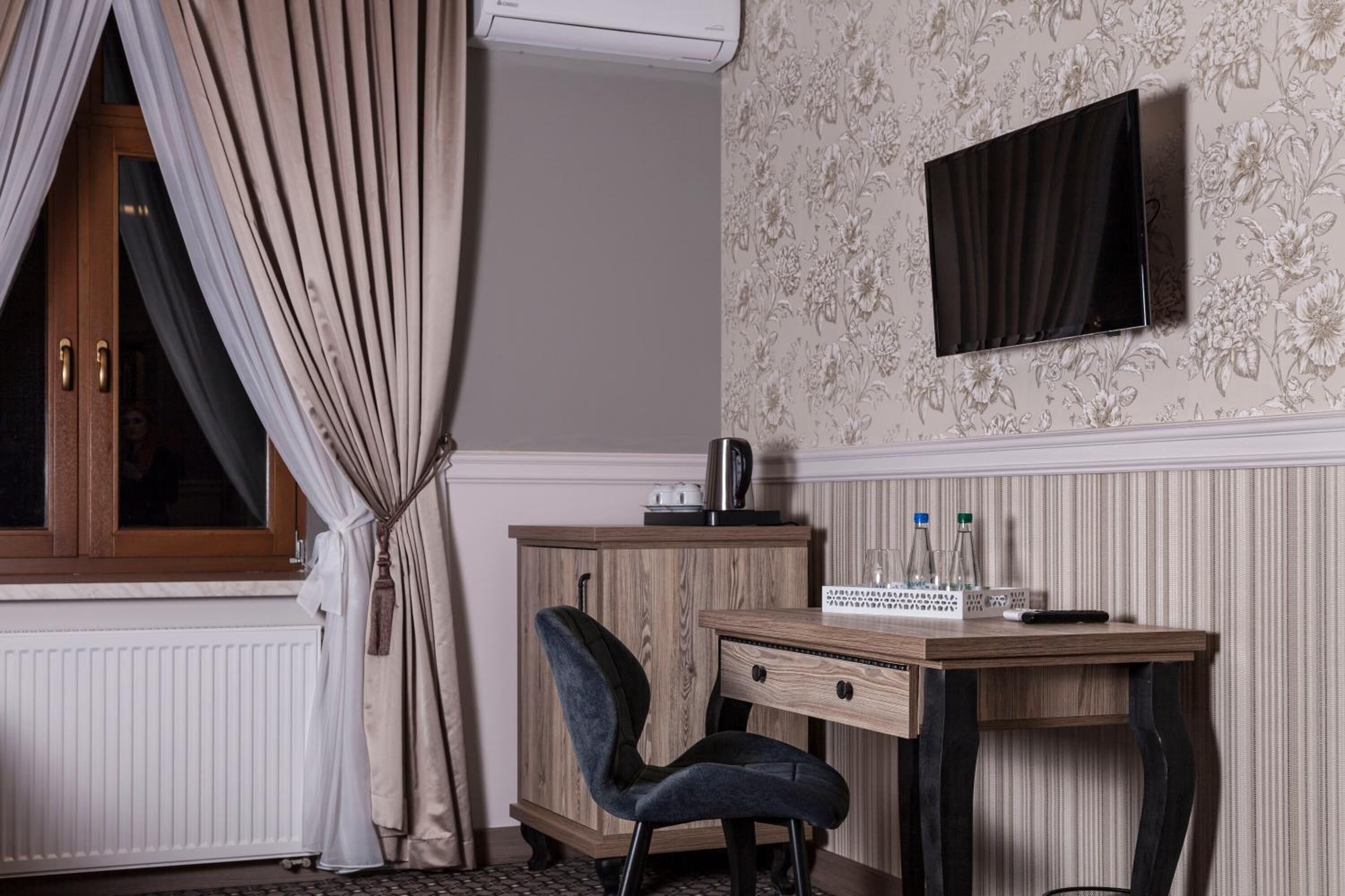A hotel room with a wooden desk, a chair, a television mounted on the wall, and a window with curtains.