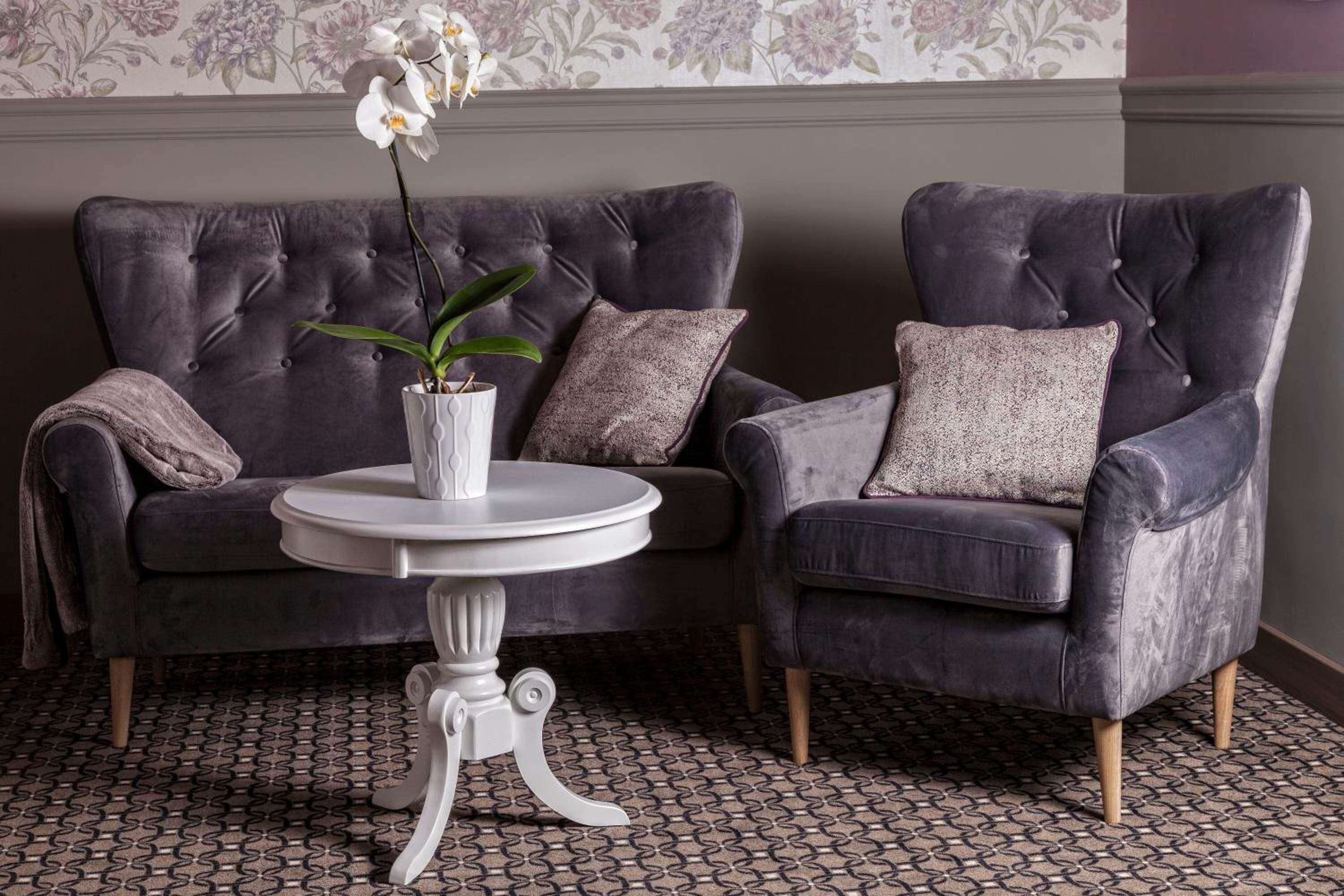A grey upholstered sofa with a white vase and orchid, a round white table, and patterned floor tiles.