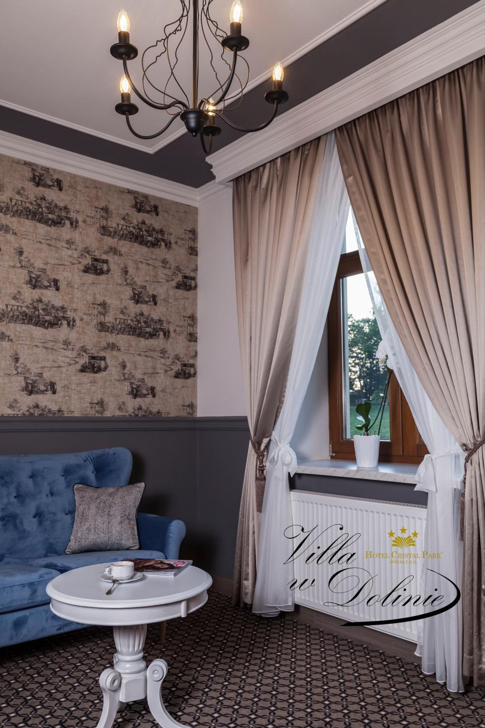 Una habitación con un sofá azul, una mesa blanca con una taza y un libro, un candelabro, un ventanal con cortinas y un radiador.