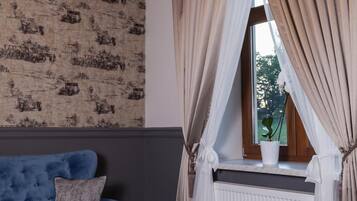 Una habitación con un sofá azul, una mesa blanca con una taza y un libro, un candelabro, un ventanal con cortinas y un radiador.