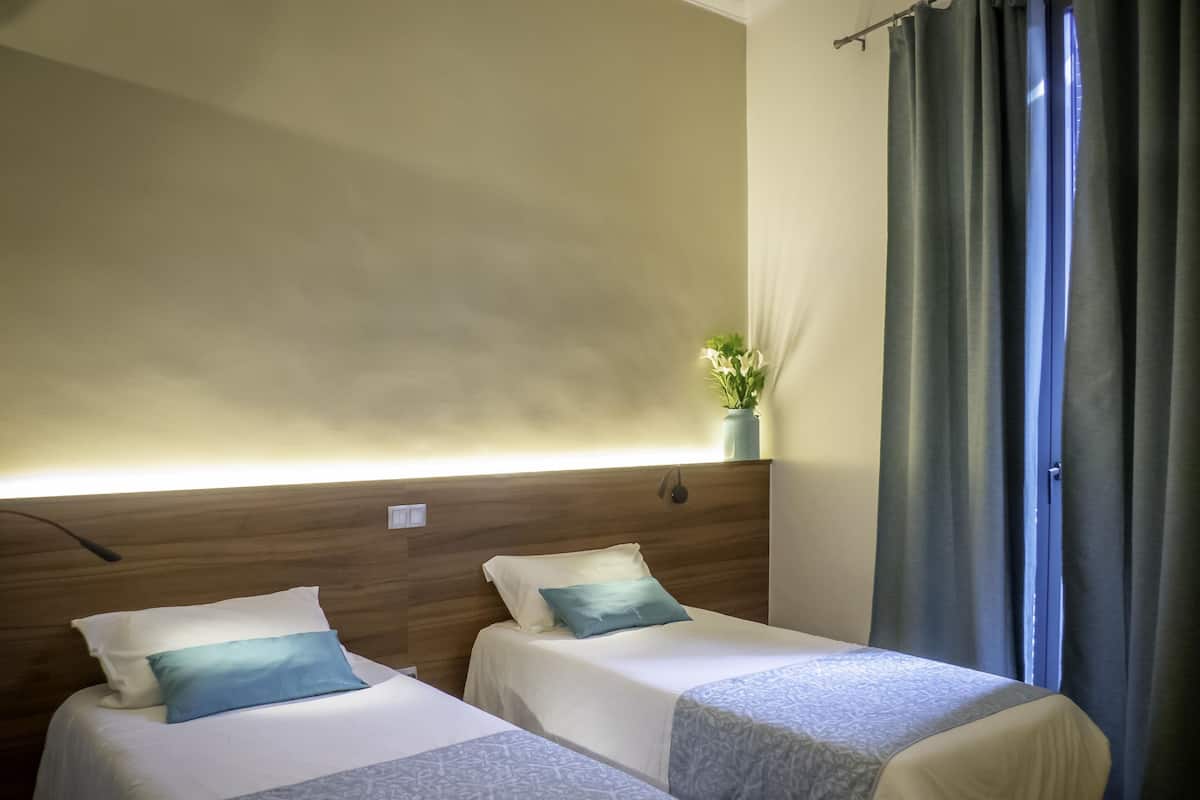 Quarto de hotel com duas camas, mesa de cabeceira, luminária na parede e janela com cortinas.