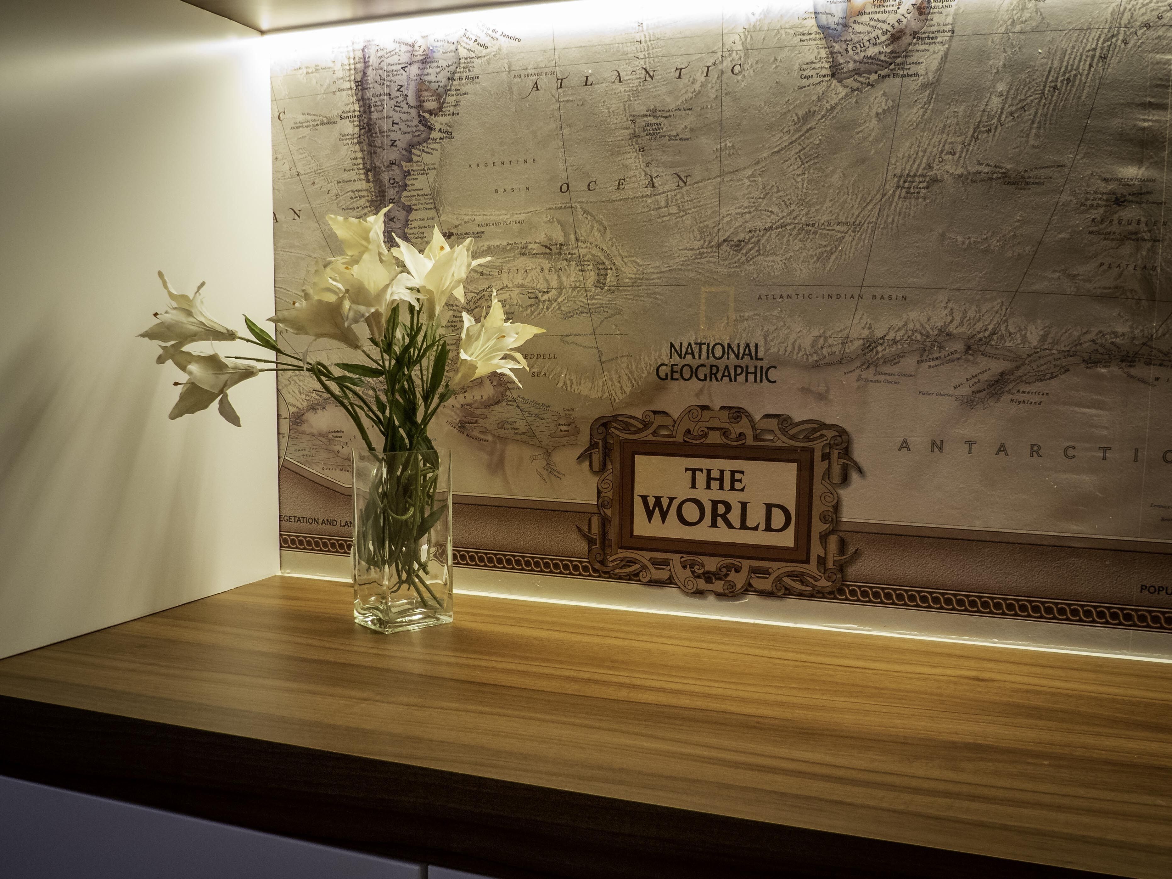 Una mesa de madera con un florero de vidrio que contiene flores blancas, frente a una pared con un mapa mundial y un letrero que dice "THE WORLD".