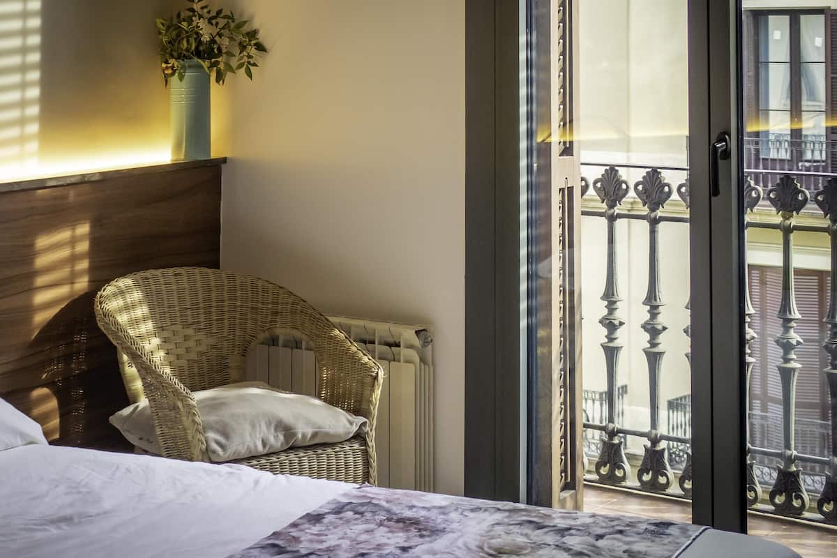 Quarto com cama, cadeira de vime, radiador, janela com vista para uma varanda e uma planta no parapeito.